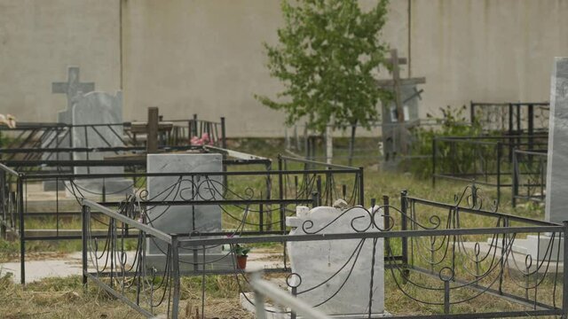 White, gray marble stone unnamed graves, cross, tombstones, with black metal fences stand in empty cemetery with green tree. View of burial place of people on background of wall. No people. Outdoors. 