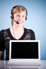 Blonde smiling girl with headset in front of  laptop. Studio photo isolated on light blue background.