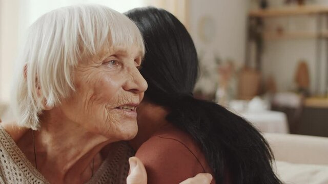 Close up shot of positive senior woman talking and hugging adult granddaughter while sitting together on sofa at home