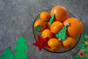 Obraz na płótnie Canvas fruits tangerines in a glass dish with New Year's decor on a gray background with cardboard Christmas trees 