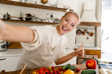 Excited woman taking selfie and gesturing thumb up while making lunch