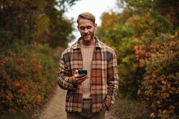 Joyful man using mobile phone while strolling in autumn forest