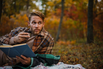 Handsome unshaven man reading book drinking tea while resting in forest