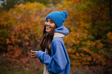 Nice happy woman in knit hat drinking tea while strolling in forest