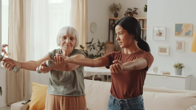 Asian female fitness instructor teaching senior woman how to do exercise with dumbbells at home