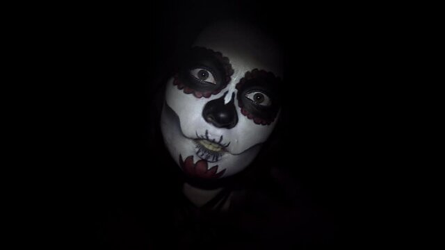 A lady with a mystic make-up in santa muerte style on black background in close-up makes frightening movements with her hands. Portrait of young woman with painted face is illuminated by a flashlight.