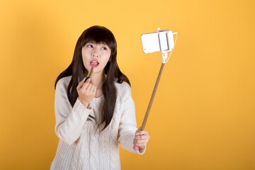 Woman vlogger holding up her phone on a selfie stick to live-streame and showing new lipstick to fans at studio on yellow background. 