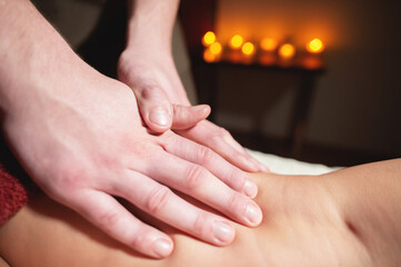 Close-up of a man's hands doing a thigh massage to a woman client in a dark room of a spa salon on the background of burning candles