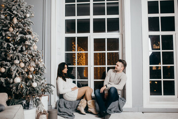 Beautiful couple sit near decorated Christmas tree, cuddling, beautiful wife with loving husband spend winter holidays together, enjoy tenderness, New Year celebration concept