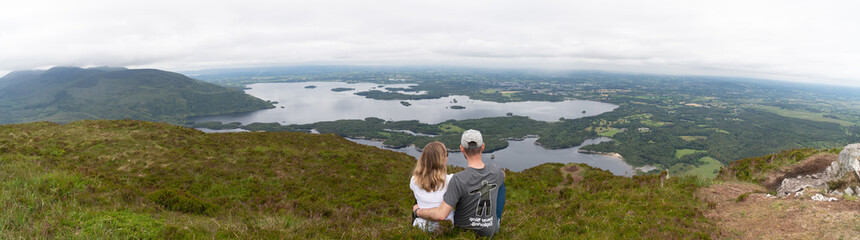 Panoramic view of the Irish countryside with trees, green vegetation and lakes with mountains and hills, cloudy day in Ireland. Couple sitting on the edge