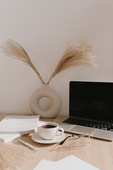 Female home office desk workspace. Blank screen laptop computer with copy space. Coffee cup, pampas grass in stylish vase on beige wooden table. Minimalist lifestyle blog mockup.