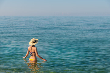 Attractive young woman in swimsuit and straw hat stands waist-deep in the calm morning sea water