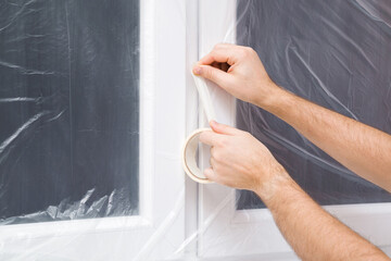 Young adult man hands using paper scotch and gluing plastic film on window frame. Closeup....