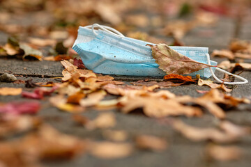 A thrown away dirty disposable face mask lying on the pavement under autumn leaves. Seen in Germany in October during corona crisis.