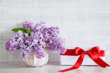 Gift box with red ribbon. Bouquet of fresh purple lilac blossoms in vase. Light gray wooden vintage background. Giving or receiving gift. Front view. Closeup.