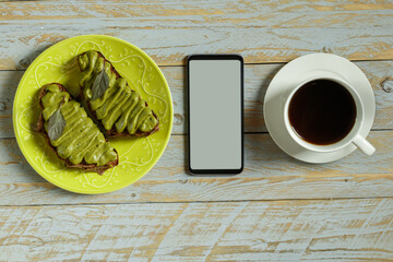 Avocado pasta with green basil, garlic and coconut cream on whole grain bread on a faience green plate next to a smartphone and a cup of coffee on a table made of stained shabby boards