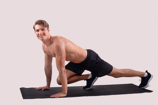 Smiling shirtless man with muscular body doing yoga or pilates on sports mat over light studio background