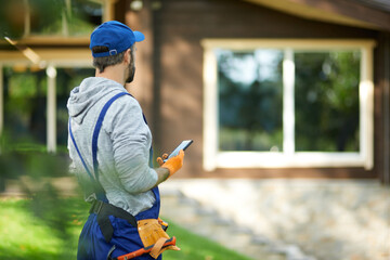 Young male builder in uniform using smartphone, standing outdoors while working at cottage...