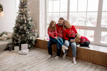 Young Caucasian beautiful family mom and dad are sitting embracing with their son, daughter and dog on the windowsill of the house among the Christmas decorations. Family portrait at home 