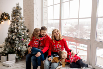 Young Caucasian beautiful family mom and dad are sitting embracing with their son, daughter and dog on the windowsill of the house among the Christmas decorations. Family portrait at home
