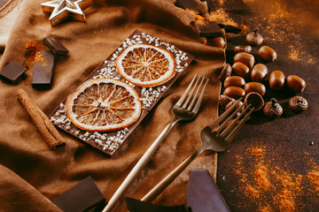 Composition of bars and pieces of different dark chocolate with orange slices, spices on a brown background close
