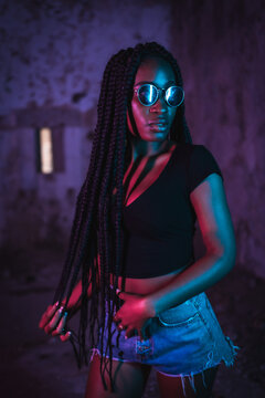 Lifestyle, Portrait Of A Young Black Woman With Long Braids, Sunglasses And A Black T-shirt. Pink And Blue Neon Lights, Urban Photography Of A Trap Dancer Looking Sideways