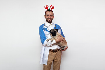 Young bearded doctor with toy deer antlers on his head holds a cute pug dog. Portrait of veterinarian doctor. New Year and Christmas concept. Copy space. Place for text