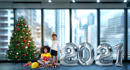 Dark skinned children have fun with metallic silver number balloons and Christmas trees, 2021 Happy new year,  against building background