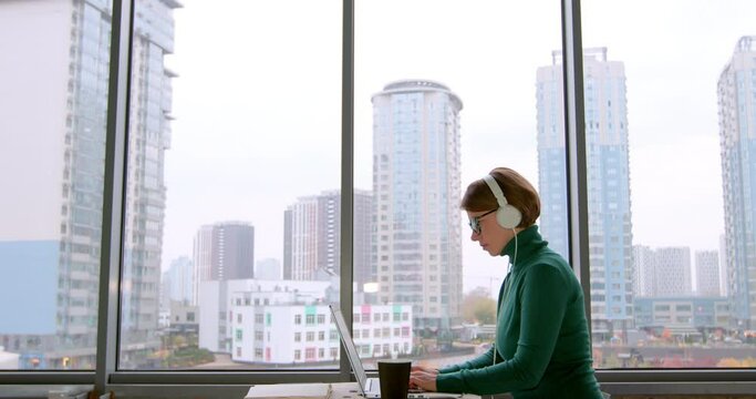 Business woman working on a laptop in headphones in an online conference. A woman in an office with large windows uses a computer. Webinar.