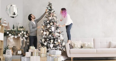 Young couple in love are helping each other hanging toys. New Year, Christmas, Xmas, holiday and celebration concept. Happy family husband and wife is decorating Christmas tree together at home. - Powered by Adobe