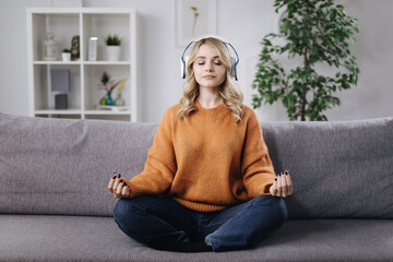 Happy young woman relaxing and meditating while using wireless headphones for enjoying favorite music. Beautiful blonde wearing knitted sweater and jeans at home.