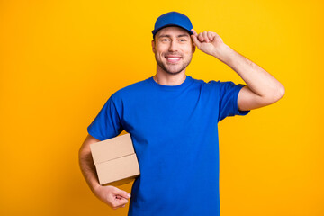 Portrait of cheerful working man arm touch cap hold carton package isolated on yellow color...