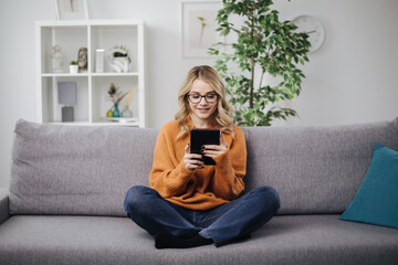 Relaxed young woman with wavy blond hair sitting on grey sofa and using digital tablet. Concept of...