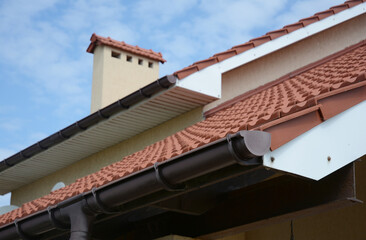 A close-up of a rain gutter with a stop end and running outlet installed correctly a few inches lower than the roofline with a blurred soffit, rooftop and chimney in the background.