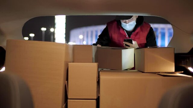 Delivery service man wearing his protective mask due to Covid-19 late nigt with a delivery item in a car. Volunteer Work in the Midst of an Epidemic. Courier with a Cardboard Box Package on a second