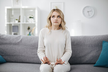 Confused young woman with wavy blond hair sitting on grey couch and looking aside. Concept of real human emotion and feelings.