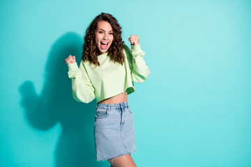 Photo of happy yell young woman raise fists dressed green sweater denim skirt isolated on turquoise color background