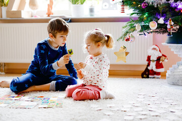 Two little chilren, cute toddler girl and school kid boy playing together card game by decorated Christmas tree. Happy healthy siblings, brother and sister having fun together. Family celebrating xmas