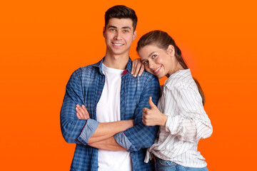 Smiling man and lady in casual clothes, look at camera, female presses to guy shoulder and shows thumb up