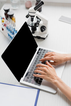 partial view of scientist typing on laptop with blank screen near microscope on desk, stock image