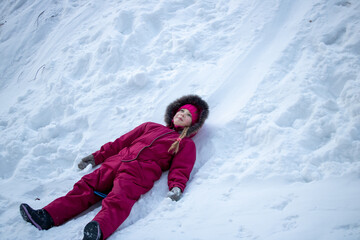 Fototapeta na wymiar A girl in a red jumpsuit rolled down a snow slide and lies on the snow and looks at the sky and smiles. The concept of the winter holidays.