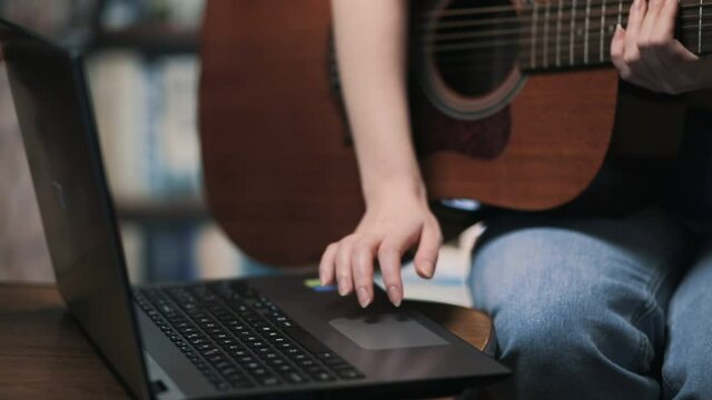 A woman is typing on a laptop, holding a guitar. Close up. Online learning to play musical instruments