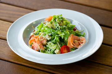 Healthy caesar salad with smoked salmon fish in a white plate on a wooden table.