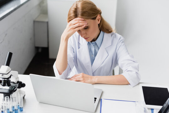 upset scientist in white coat covering face near laptop and microscope on desk, stock image