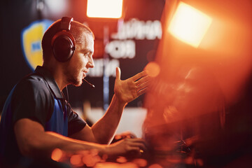 Side view of a professional male cybersport gamer wearing headphones talking by microphone with...