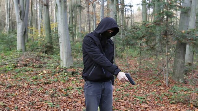A man armed with a firearm stands in the forest.A man wearing a balaclava mask finishes his victim with a pistol shot.Video contains sound.