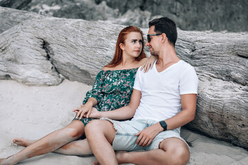 Hipster couple on the beach, girl with red hair in green floral dress and man with stubble and tattoo "Timon" on his arm, sitting on the old big bare tree without leaves. Phuket. Thailand