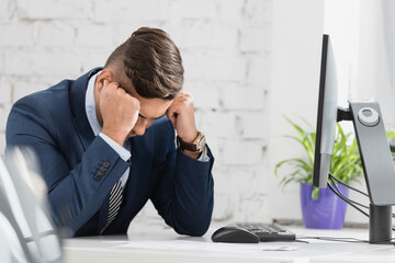  businessman with hands near head, sitting near computer at workplace on blurred foreground