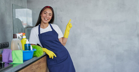 Cheerful young woman, cleaning lady in protective gloves smiling at camera, pointing up while...