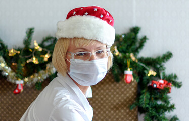 Smiling middle - aged woman in a medical mask and Santa hat. Remote congratulations on Christmas. Pandemic.